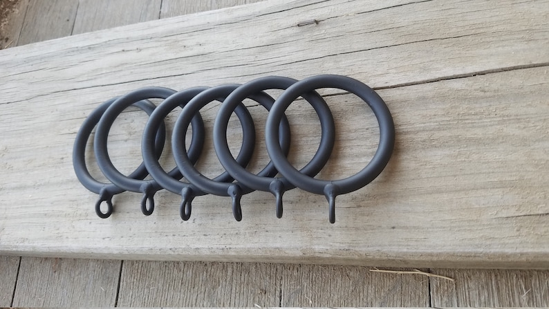 May include: Six black metal curtain rings with a hook on each ring. The rings are arranged in a row on a wooden surface.