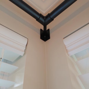May include: A black metal curtain rod set installed in a corner. The rod is composed of two straight sections joined at a 90-degree angle. The rod is mounted on a wall with a neutral color, and the window blinds are visible.
