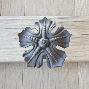 May include: A decorative, cast metal door pull or cabinet knob in a stylized leaf design. The gray-toned hardware is mounted on a light-colored wooden surface. The leaf has a central spiral and radiating, textured segments.