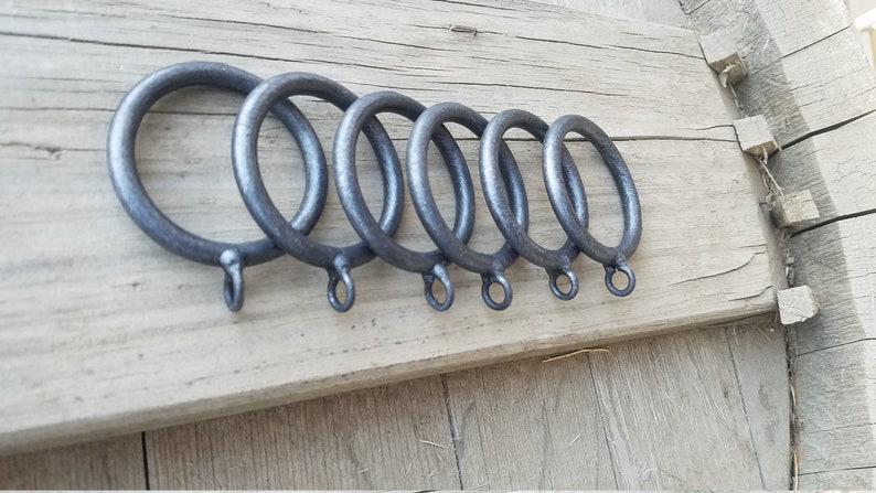 May include: Six dark gray metal curtain rings of varying sizes are arranged on a weathered wooden surface. Each ring has a small loop at the bottom. The rings are likely for hanging curtains or drapes.