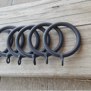 May include: Six dark gray metal curtain rings with a simple, round design. Each ring has a small hook at the bottom for attaching to curtains. The rings are arranged on a weathered wooden surface.