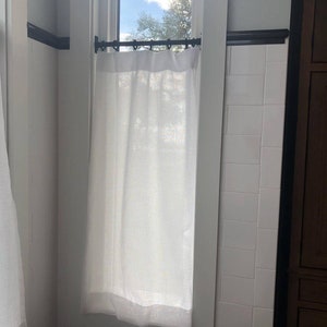 May include: A white linen curtain hangs from a black metal rod in front of a window. The window is framed in white trim and is set against a white tiled wall.