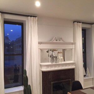 May include: Interior shot of a living room with white curtains framing two windows. A fireplace with a decorative mantel and deer figurines is centered between the windows. A cactus is in front of the window.