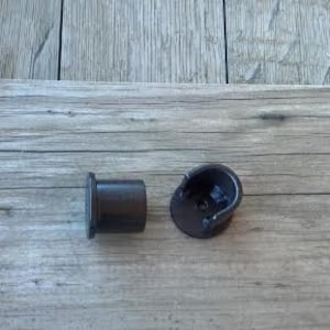 May include: Two dark brown, cylindrical metal hardware components. One piece is a solid, flat-ended cylinder, while the other is a matching cylindrical bracket with an open, U-shaped top section and a central screw hole for mounting purposes.