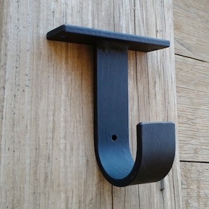 May include: Black metal curtain rod bracket with a J-shaped hook. The bracket is mounted to a wooden surface.