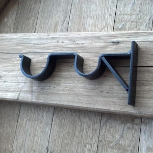 May include: Black metal curtain rod bracket with two curved arms and a triangular support. The bracket is mounted on a wooden surface.