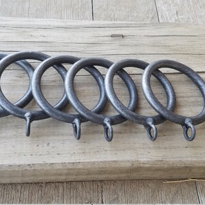 May include: Five dark gray metal curtain rings with attached hooks are arranged in a row. The rings are circular and have a slightly textured surface. They rest on a weathered, light brown wooden surface.