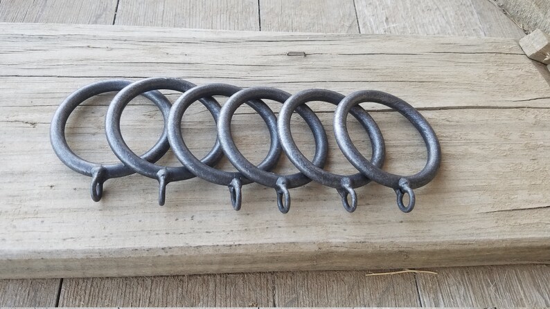May include: Five dark gray metal curtain rings with attached hooks are arranged in a row. The rings are circular and have a slightly textured surface. They rest on a weathered, light brown wooden surface.