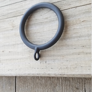 May include: A matte black curtain ring with a circular design and an attached loop. The ring is made of metal and is set against a weathered, light-colored wooden surface. This hardware is suitable for curtains and draperies.