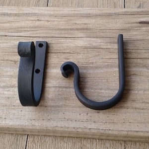 May include: Two black, hand-forged iron hooks. One is a wall-mounted hook with a curved top and two mounting holes. The other is a J-shaped hook with a decorative curl. Both are on a weathered wooden surface.