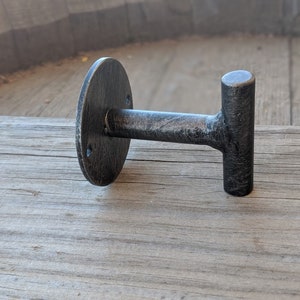May include: A black metal T-shaped hook with a round mounting plate. The hook is attached to a wooden surface.