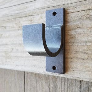 May include: A metal curtain rod bracket with a U-shaped hook. The bracket is painted a dark gray color and has two small holes on the back for mounting.