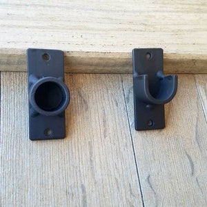May include: Two black metal curtain rod brackets mounted on a wooden wall. One bracket has a round opening and the other has a curved opening.