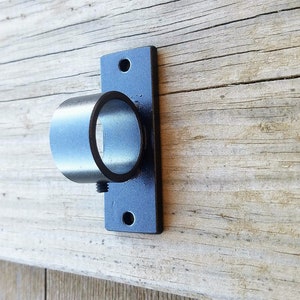 May include: Black metal bracket with a round opening for a curtain rod. The bracket is mounted on a weathered wooden surface.