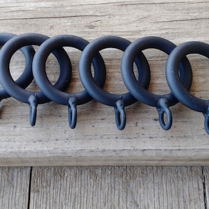 May include: Six black metal curtain rings with a hook on each ring. The rings are arranged in a row on a wooden surface.