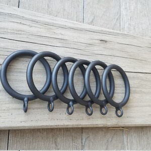 May include: Six black metal curtain rings of varying sizes, arranged in a row on a wooden surface.