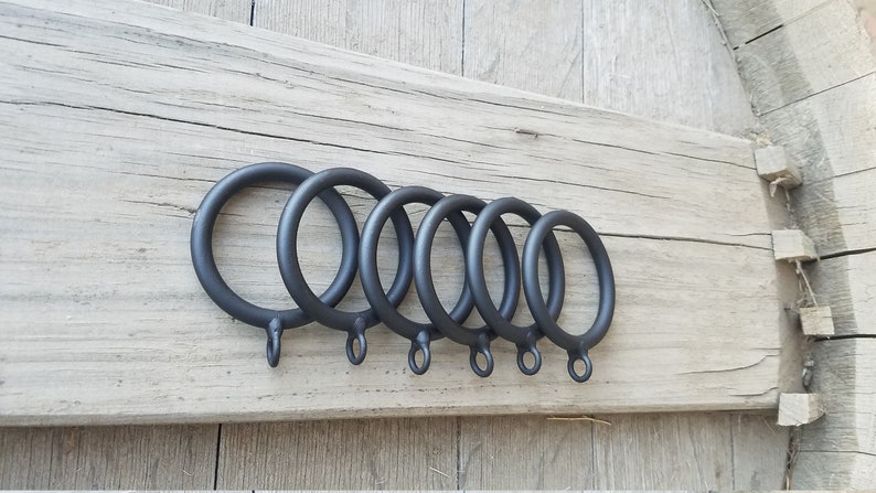May include: Six black metal curtain rings of varying sizes, arranged in a row on a wooden surface.