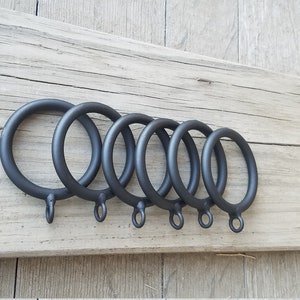 May include: Six black metal curtain rings of varying sizes are arranged on a weathered wooden surface. Each ring has a small loop at the bottom for attaching curtains. The rings are a matte black finish.