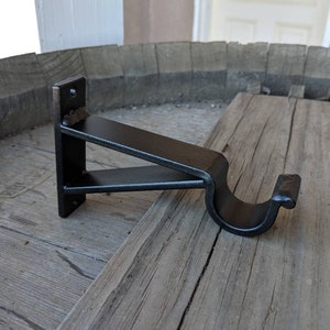 May include: Black metal curtain rod bracket with a curved arm. The bracket is mounted on a wooden surface.