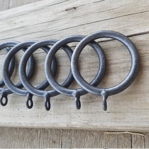 May include: Six dark gray metal curtain rings with a textured finish are arranged on a weathered wooden surface. Each ring has a small loop at the bottom for attaching curtains. The rings are evenly spaced.