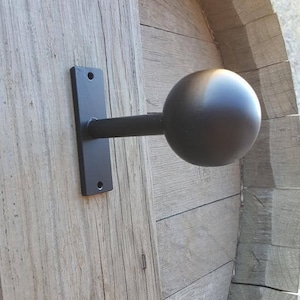 May include: A black metal curtain rod finial with a spherical end. The finial is mounted on a rectangular plate with two screw holes, attached to a weathered wooden surface. The rod extends horizontally.