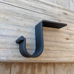 May include: Black metal shelf bracket with a curved arm and a straight arm. The bracket is mounted to a wooden surface.