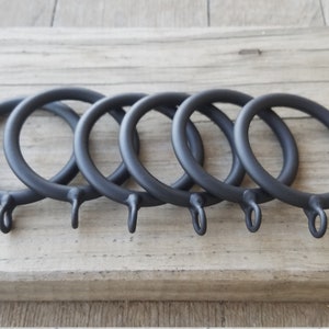 May include: Five matte black curtain rings with a simple, circular design. Each ring has a small loop at the bottom for attaching to a curtain. The rings are arranged on a light-colored wooden surface.