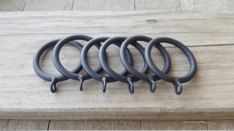May include: Five matte black curtain rings with a simple, circular design. Each ring has a small loop at the bottom for attaching to a curtain. The rings are arranged on a light-colored wooden surface.