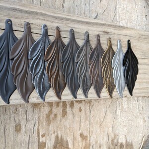 May include: Ten metal leaf charms in various finishes, including black, brown, silver, copper, gold, and grey. The charms are arranged in a row on a wooden surface.