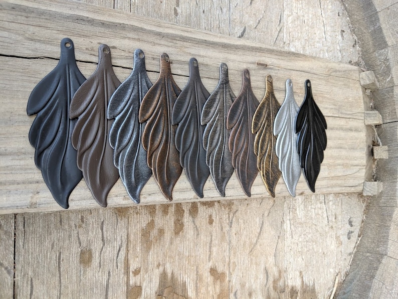 May include: Ten metal leaf charms in various finishes, including black, brown, silver, copper, gold, and grey. The charms are arranged in a row on a wooden surface.