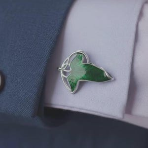 Puede incluir: Un gemelo de metal plateado con un diseño de hoja verde estilizada con detalles de nervaduras visibles, asegurado a través del ojal del puño de una camisa clara, parcialmente cubierto por la manga de una chaqueta de traje azul oscuro texturizada.