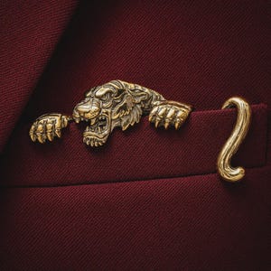 May include: A gold-toned metal brooch shaped like a tiger head with extended paws and a curved tail. The tiger is depicted with an open mouth showing teeth, and the piece is attached to the edge of a dark red fabric pocket, creating a three-dimensional effect.
