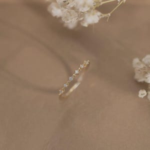 May include: A thin gold ring featuring a delicate band adorned with a series of small, clear, round-cut stones set in a row. The metal has a polished finish, and the stones are spaced evenly along the top portion of the circular band structure.