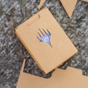 May include: A brown cardboard box with a die-cut logo featuring a stylized hand with a purple and blue design. The box is rectangular with a tab closure and is surrounded by cut cardboard pieces.
