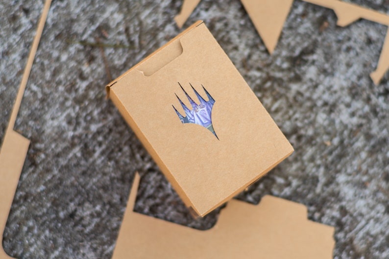 May include: A brown cardboard box with a die-cut logo featuring a stylized hand with a purple and blue design. The box is rectangular with a tab closure and is surrounded by cut cardboard pieces.