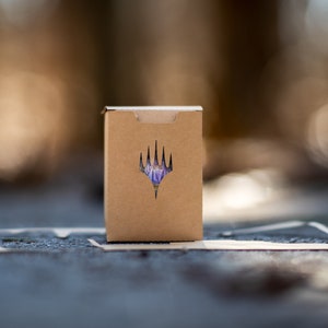 May include: A small, brown cardboard box featuring a die-cut logo on the front. The logo is a stylized, purple and white emblem. The box is on a textured surface with a blurred background.