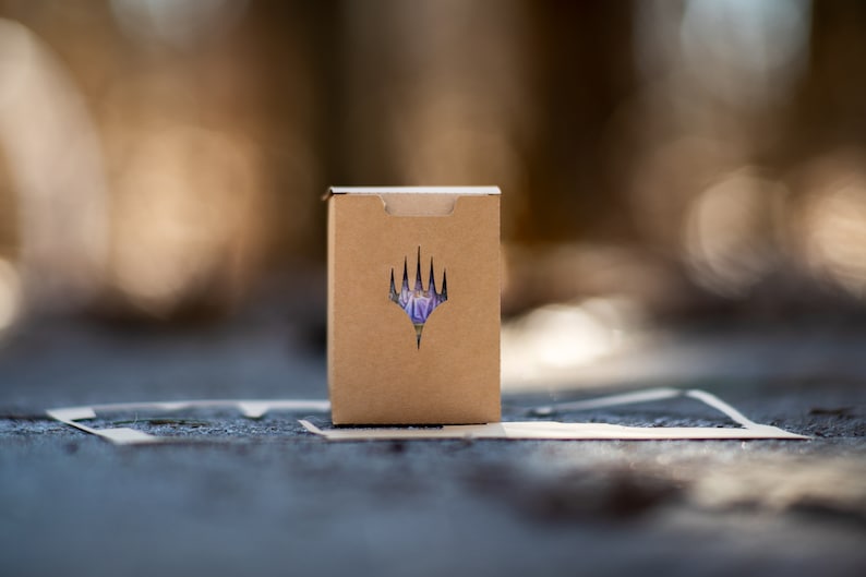May include: A small, brown cardboard box featuring a die-cut logo on the front. The logo is a stylized, purple and white emblem. The box is on a textured surface with a blurred background.