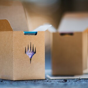 May include: Two open brown cardboard boxes, each with a die-cut logo on the front. The logo is a stylized, multi-pointed shape. The boxes are on a textured surface. The boxes are designed to hold a collectible card game.