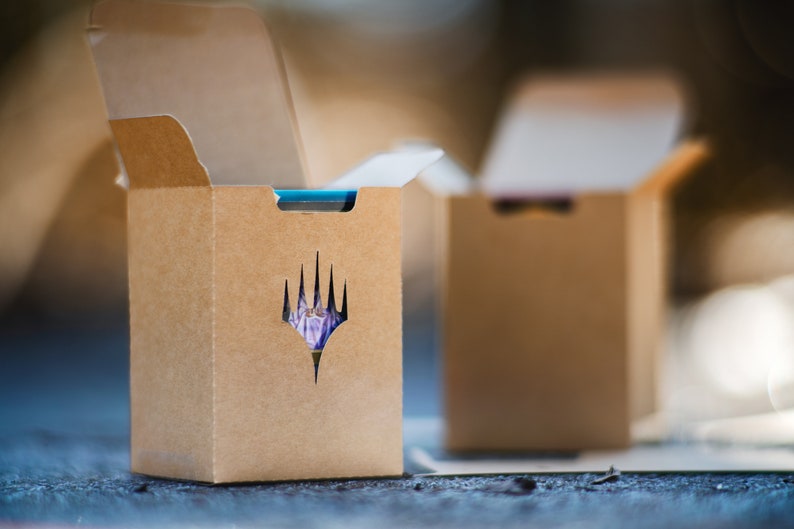 May include: Two open brown cardboard boxes, each with a die-cut logo on the front. The logo is a stylized, multi-pointed shape. The boxes are on a textured surface. The boxes are designed to hold a collectible card game.