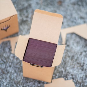 May include: Kraft card boxes, one open to show a stack of burgundy cards. The boxes have a die-cut design on the front and are designed to hold cards. The boxes are on a textured surface.