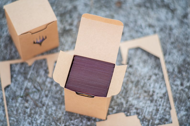 May include: Kraft card boxes, one open to show a stack of burgundy cards. The boxes have a die-cut design on the front and are designed to hold cards. The boxes are on a textured surface.