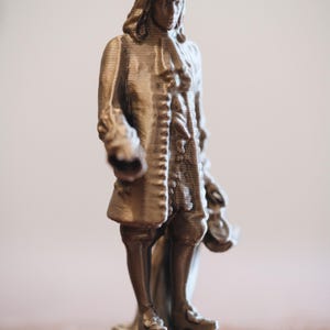May include: A bronze-colored figurine of a man in 18th-century attire, including a wide-brimmed hat and long coat. The figure is standing on a small base, and the details of the clothing and face are visible.