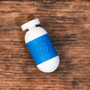May include: A white and blue bottle with a blue band featuring a circular design. The bottle has a rounded shape and a white cap. The object is set against a wooden background.