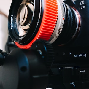 Könnte beinhalten: Nahaufnahme eines schwarzen Kameraobjektivs mit einem roten Zahnradring. Das Objektiv ist mit "Konica Hexanon" beschriftet und trägt die Nummer "7204071". Das Kameragehäuse ist schwarz und trägt das "SmallRig"-Logo.