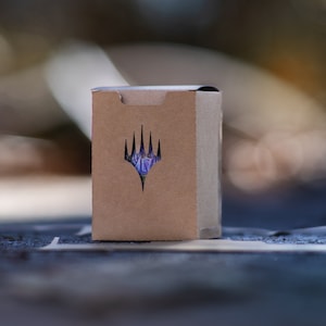 May include: A brown cardboard box featuring a die-cut logo of a stylized crown. The box is closed and likely contains a collectible card game. The background is blurred, suggesting an outdoor setting.