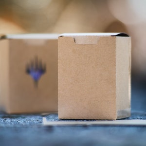 May include: Two small, rectangular, brown cardboard boxes. The box in the background has a blurred blue and purple graphic. The boxes are on a blue surface with white lines. The boxes are in focus, with a blurred background.