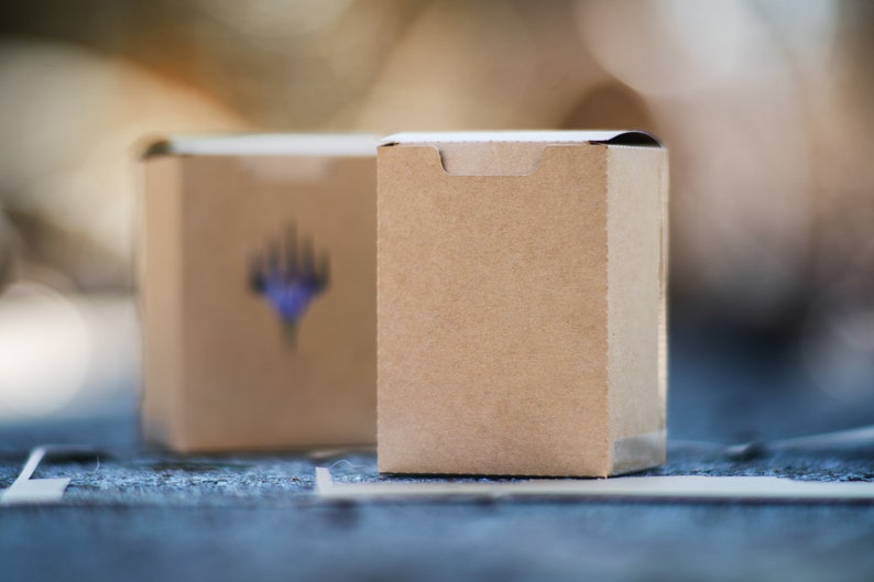 May include: Two small, rectangular, brown cardboard boxes. The box in the background has a blurred blue and purple graphic. The boxes are on a blue surface with white lines. The boxes are in focus, with a blurred background.