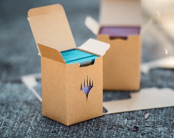 Könnte beinhalten: Zwei offene Pappschachteln, eine im Fokus, gefüllt mit türkisfarbenen und lila Spielkarten. Die vordere Box hat ein ausgestanztes Symbol. Die Boxen stehen auf einer strukturierten Oberfläche, was auf eine Tischplatte hindeutet. Kartenspeicherboxen.
