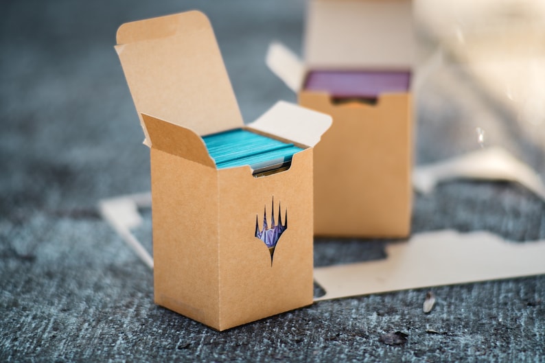 May include: Two open cardboard boxes, one in focus, filled with turquoise and purple playing cards. The front box has a die-cut symbol. The boxes are on a textured surface, suggesting a tabletop setting. The boxes are designed for card storage.