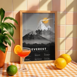 May include: A framed poster featuring a stylized grayscale illustration of Mount Everest with a yellow sun, text reading EVEREST, and various geographical facts. The print is displayed in a light wood frame on a checkered surface.